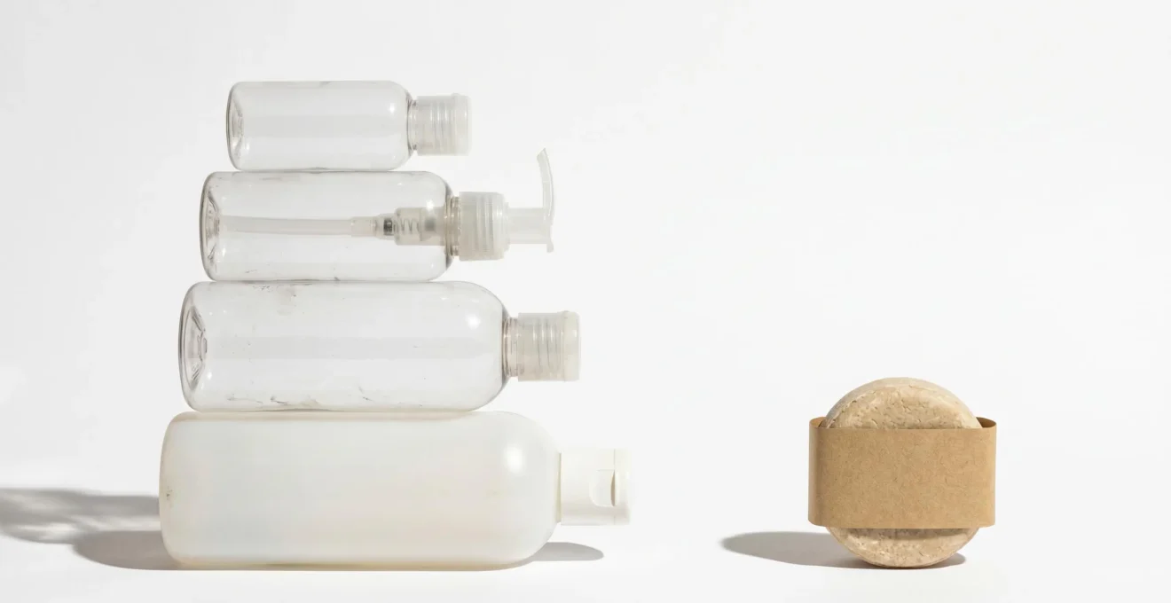 Comparaison visuelle emballages cosmétiques : empilement de flacons plastiques vides face à un shampoing solide avec emballage kraft minimal sur fond blanc
